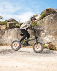 Person riding a small OTIDA R7 MAX Dual Motor Electric Bike on rocky terrain with a clear sky.