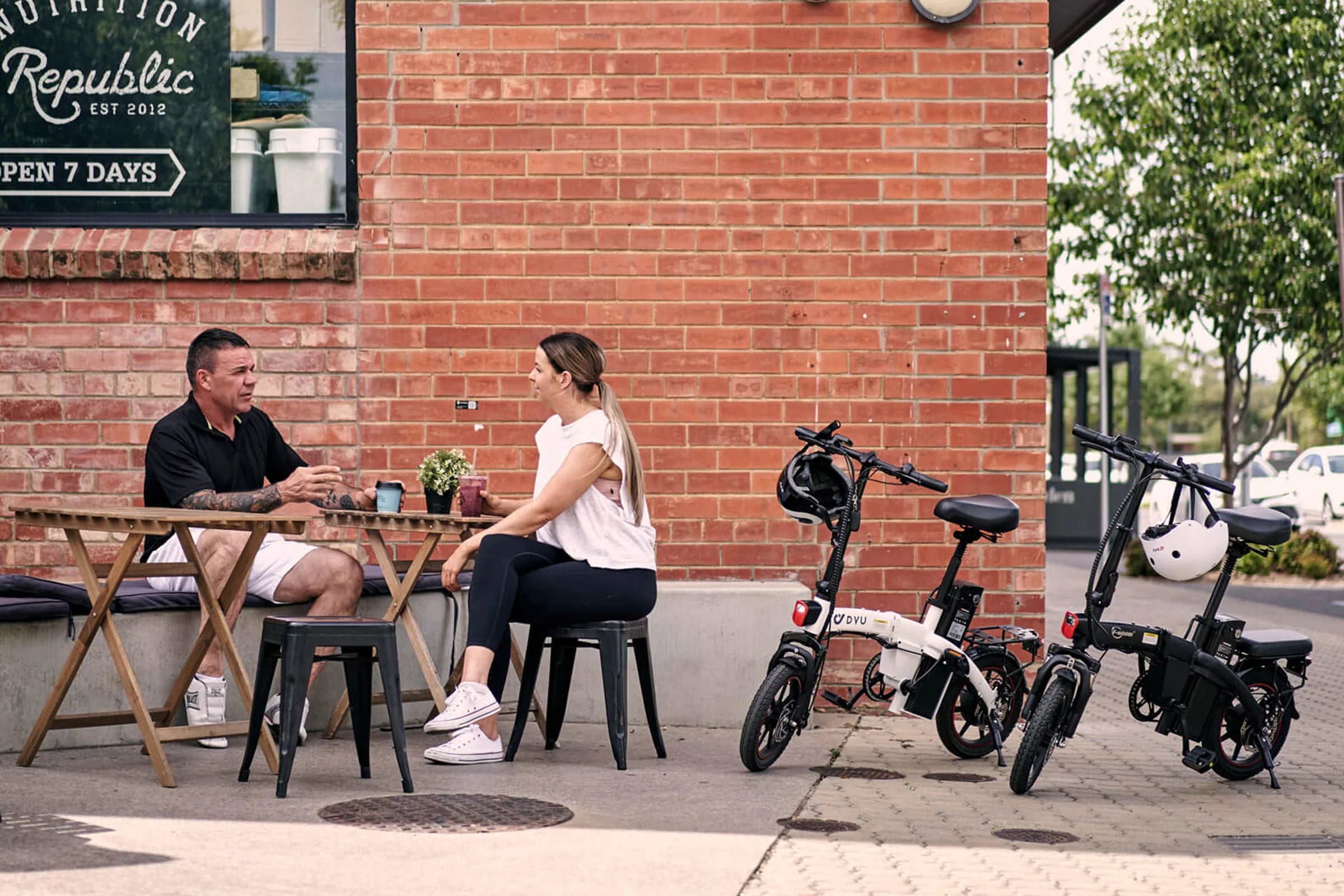 Two DYU A5 Upgraded Folding Electric bikes parked on a sidewalk next to a couple sitting at an outdoor table.