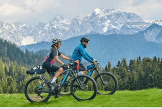 Two electric mountain bikes ridden by cyclists on grassy trail with mountains in background, side view