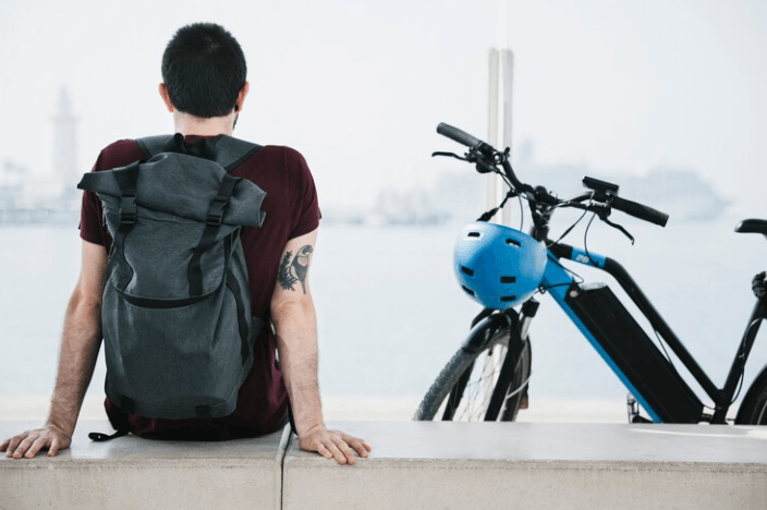 Man with backpack sitting by blue electric bike and helmet near waterfront, Pogo Cycles